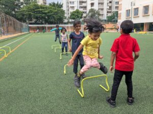 Children practice jumping over yellow hurdles on a green outdoor field, with an apartment building and trees in the background.