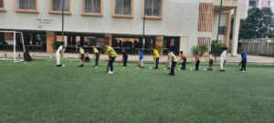 A line of children practicing cricket drills on an artificial turf field, with a building and goalpost in the background.