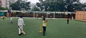 Children are playing cricket on a green artificial turf field at Akshara International School, with buildings and trees in the background.