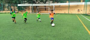 Kids in green and orange sports uniforms playing soccer on a green field, with a goalpost and fenced area in the background.
