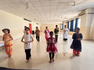 Young girls in colorful traditional outfits performing a dance routine indoors on a polished floor with ceiling fans overhead.