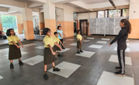 Children in yellow uniforms exercising indoors on a tiled floor, guided by an adult in a black tracksuit.