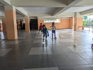 Young children in roller skates practicing indoors, moving around cones on a tiled floor under a covered area.